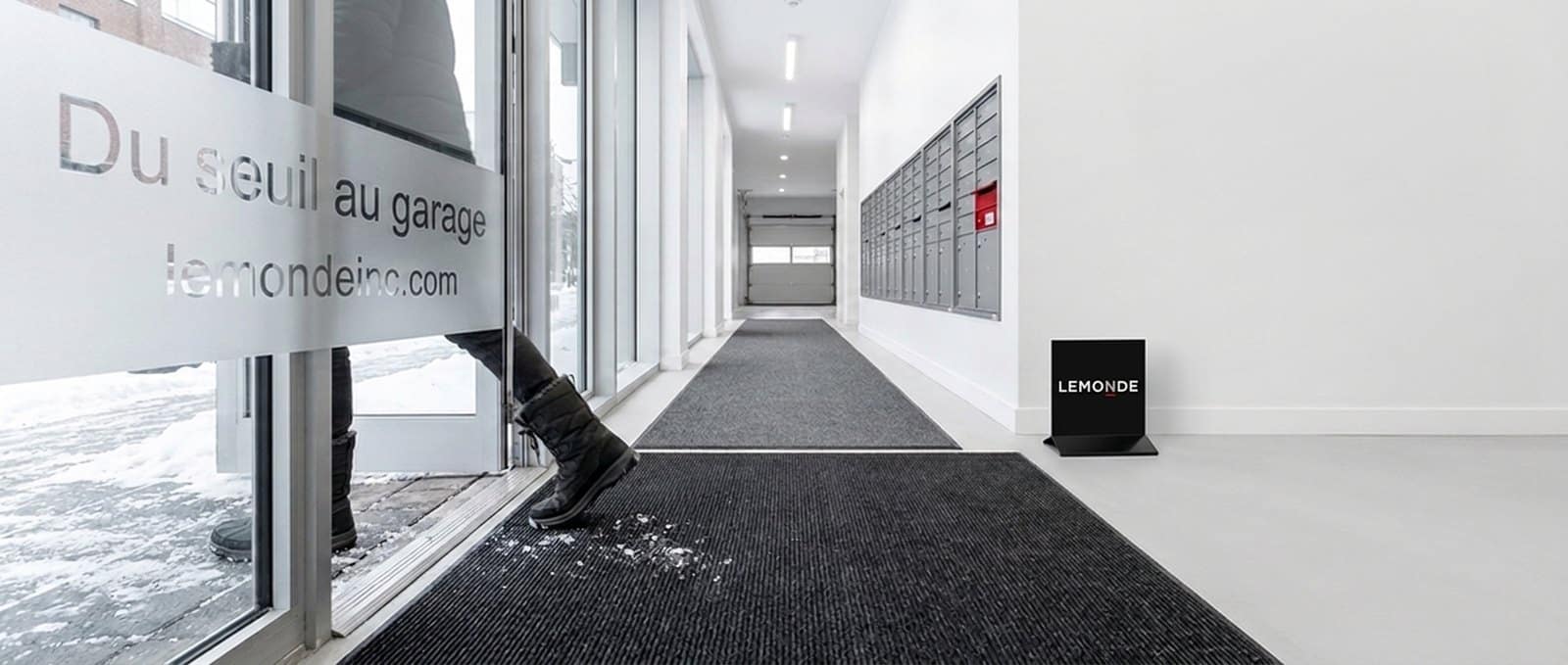 Tapis d’entrée commercial gratte-pieds puis absorbant posé du seuil jusqu’au garage dans le hall d’un immeuble, le long des boîtes aux lettres
